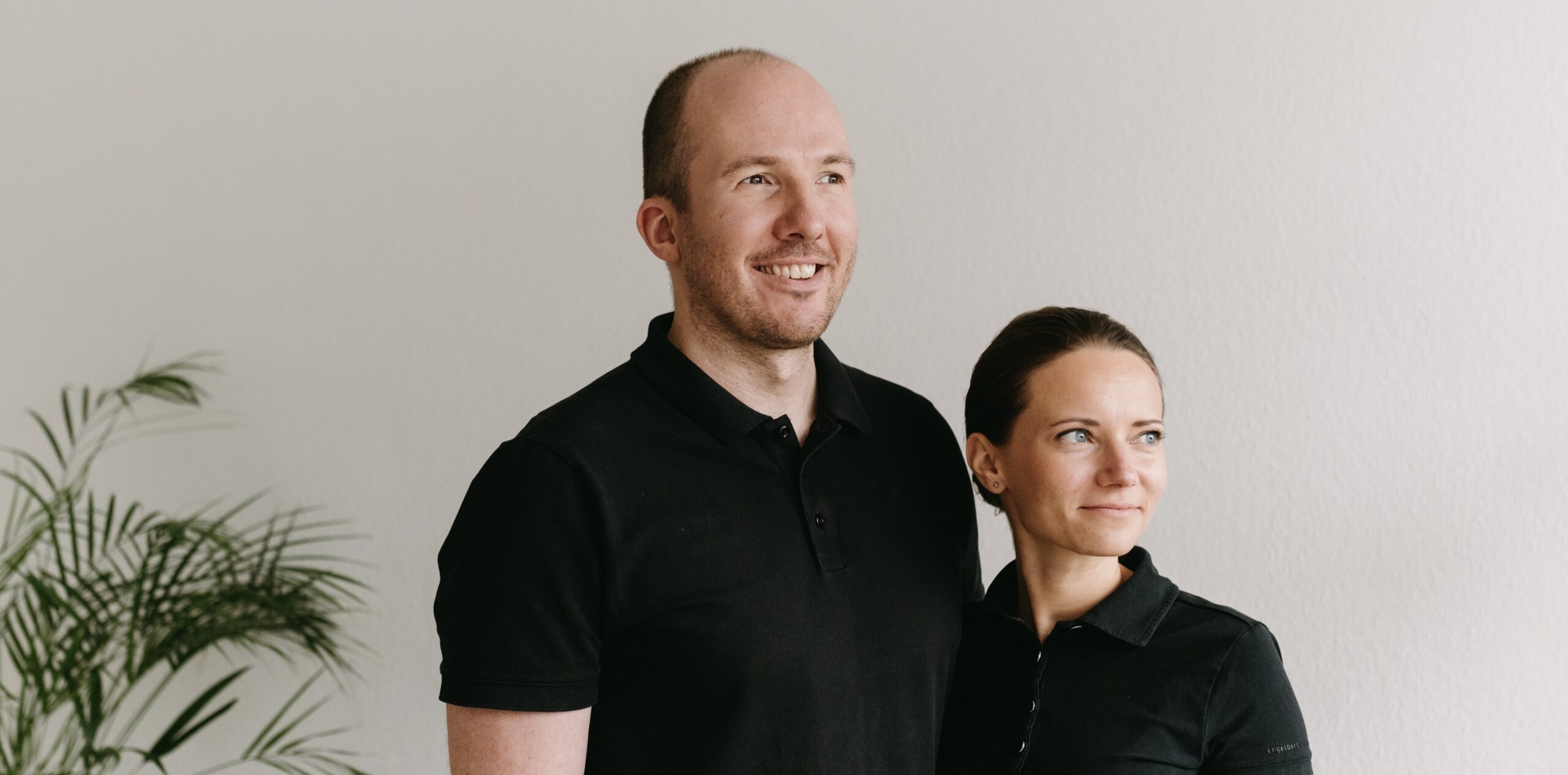 Praxis 55 - Caroline & Sven - Physiotherapie Die Physiotherapeuten der Praxis 55, Sven Kucharzewski und Caroline Siegert-Kucharzewski, in schwarzen Polo-Shirts schauen rechts aus dem Bild heraus